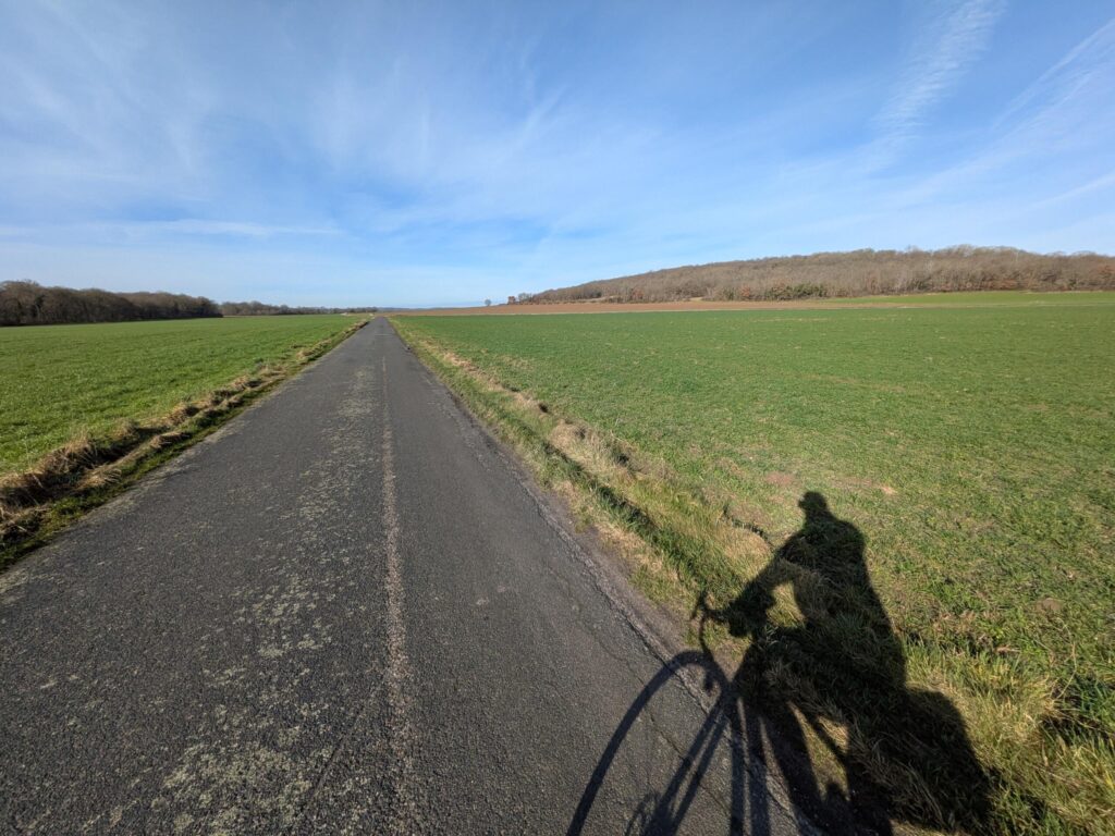 En plein jour une route de campagne avec quelques colinnes et l'ombre d'un cycliste qui prend la photo au premier plan sur la droite