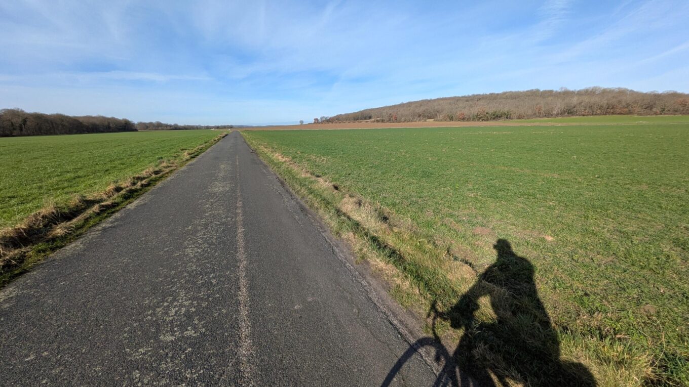 En plein jour une route de campagne avec quelques colinnes et l'ombre d'un cycliste qui prend la photo au premier plan sur la droite