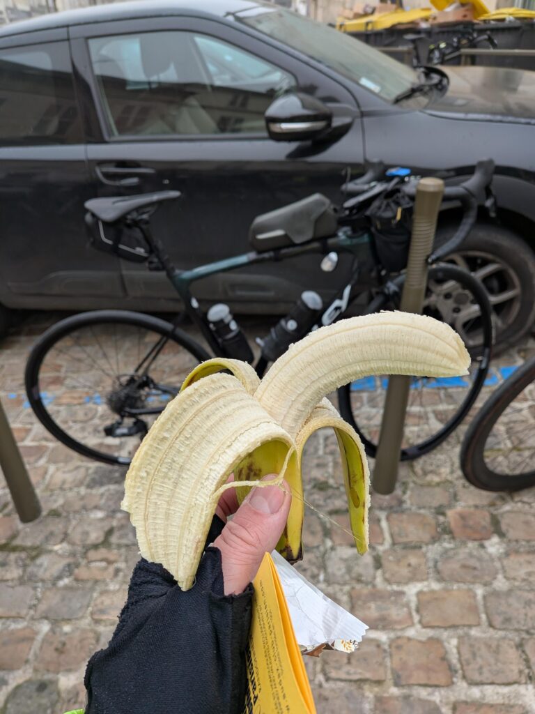 au premier plan une personne tient une banane épluchée, derrière un vélo type course appuyé sur un poteau