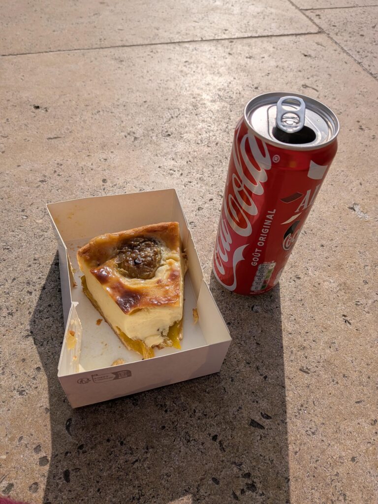 Un flan croqué dans une petite boite en carton et une cannette de coca ouverte, le tout posé au sol ensoleillé