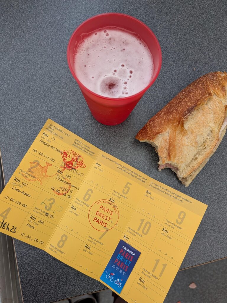 sur une table, un verre de bière, un sandwich croqué et une carte jaune avec plusieurs numéros qui sont marqués de tampons et d'heure de passage