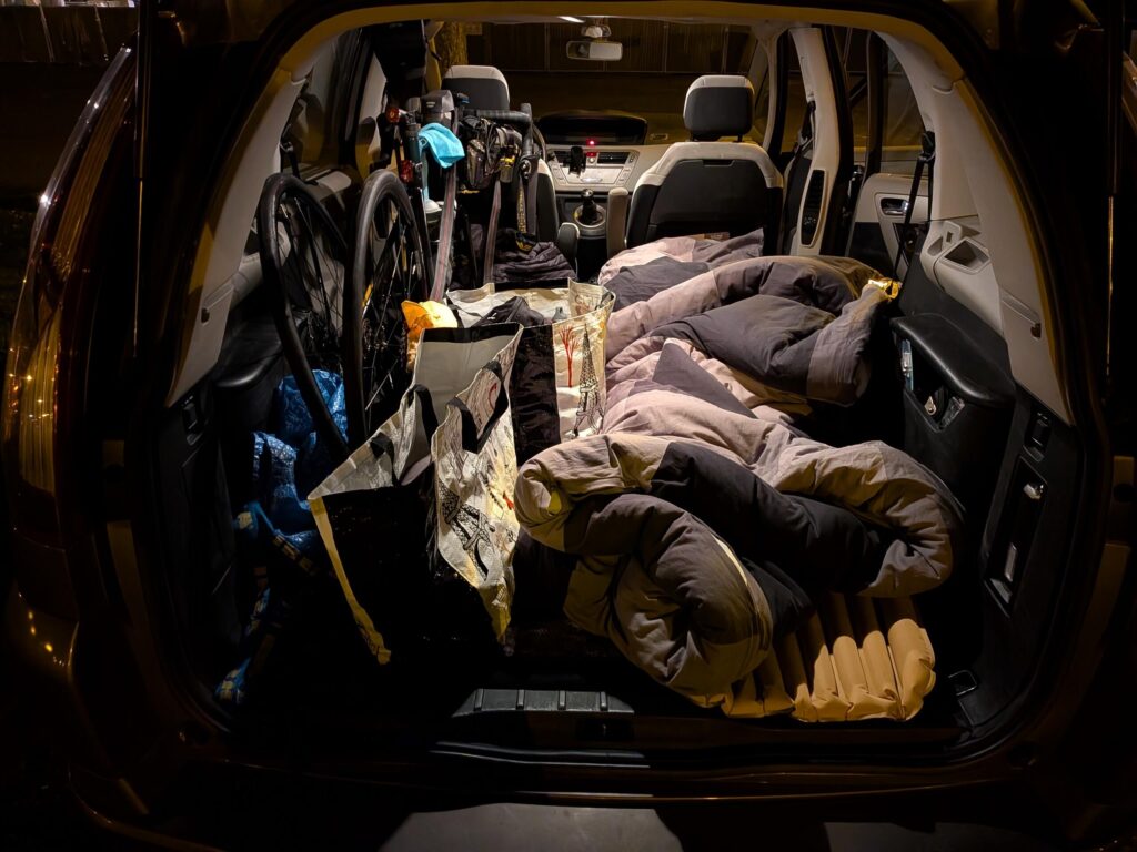 De nuit, un coffre de voiture de type monospace grand ouvert éclairé par les lampes de la voiture. Dedans un vélo, des sacs, un matelas et une couette