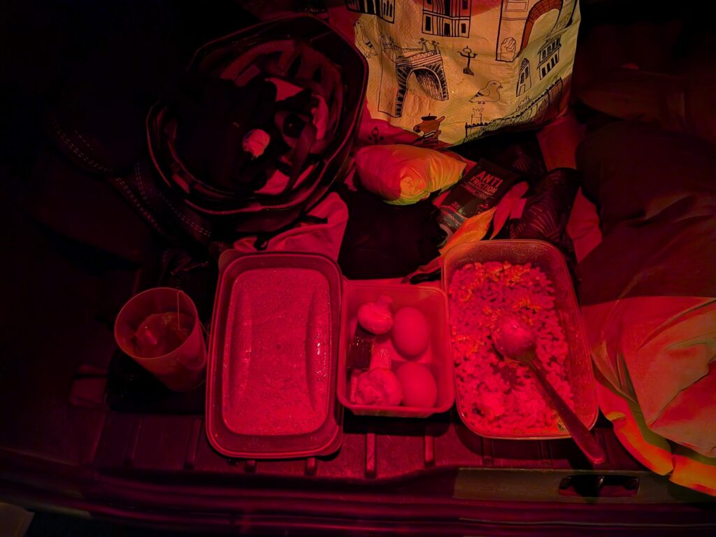 De nuit, éclairage rouge d'une frontale, une boite avec 2 œufs durs, une autre avec des pâtes, une éco-cup avec un thé. Autour entre autres un casque de vélo et un tube de crème anti friction