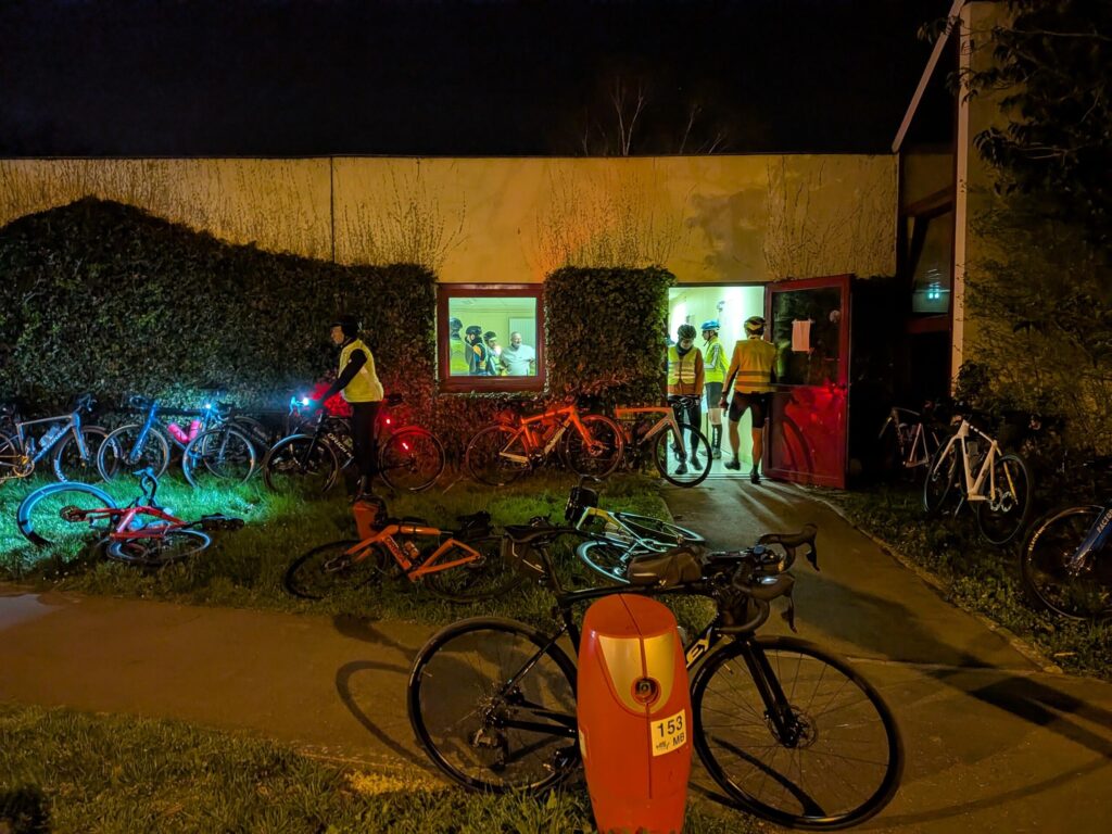 De nuit, devant l'entrée d'un local avec quelques cyclistes portant tous un gilet jaune et de nombreux vélos posés sur les murs ou sur l'herbe