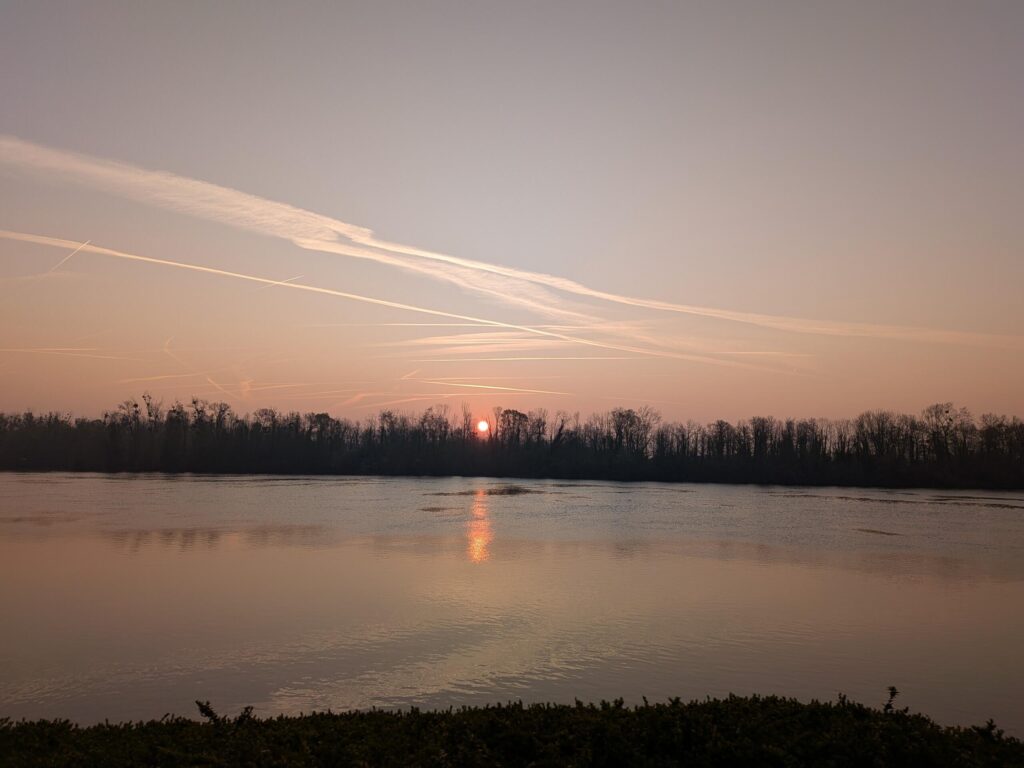 Levé du jour sur un fleuve, ciel orangé et arbres à contre jour. Au milieu le soleil sort d'entre les arbres