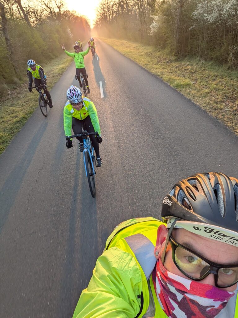 Au petit matin un cycliste fait un selfie et derrière lui une petite dizaine d'autres cyclistes. La route est étroite, dans les bois. Au fond le soleil se lève et un peu de brume