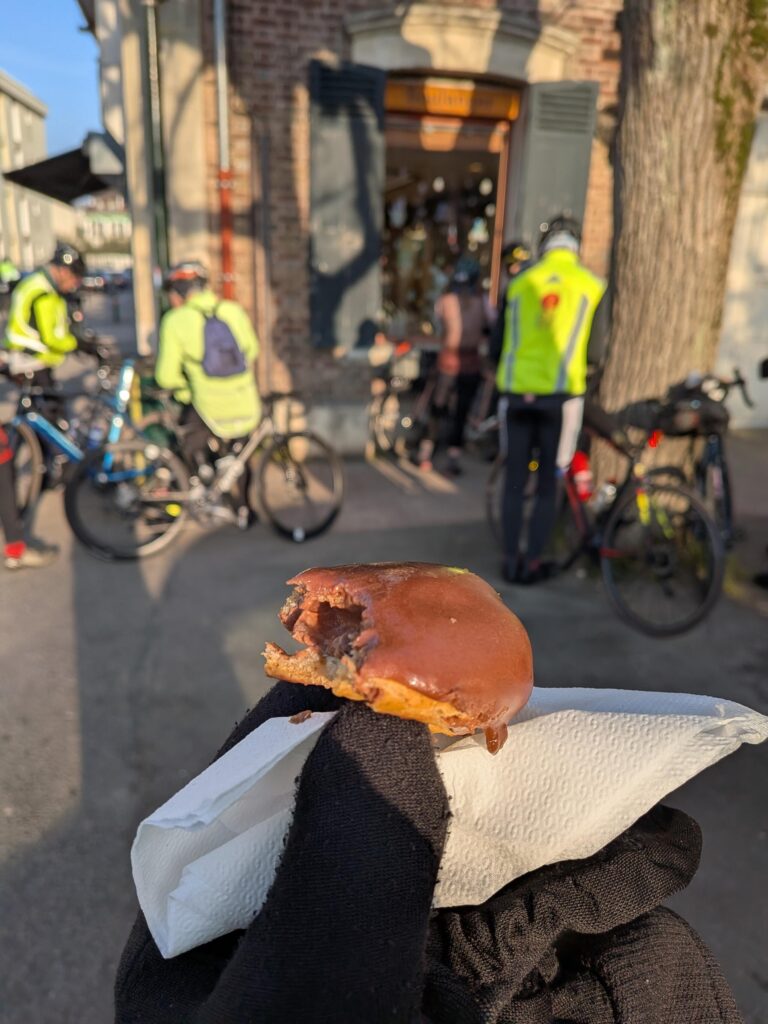 À la sortie d'une boulangerie, une personne tient le reste d'un éclair au chocolat dans sa main en gros plan. Au fond plusieurs cyclistes avec des gilets jaunes se reposent ou croquent un morceau