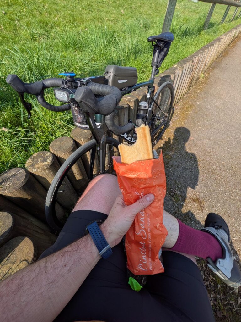 Vue subjective à la première personne d'un cycliste avec un sandwich croqué à la main, son vélo posé en arrière plan