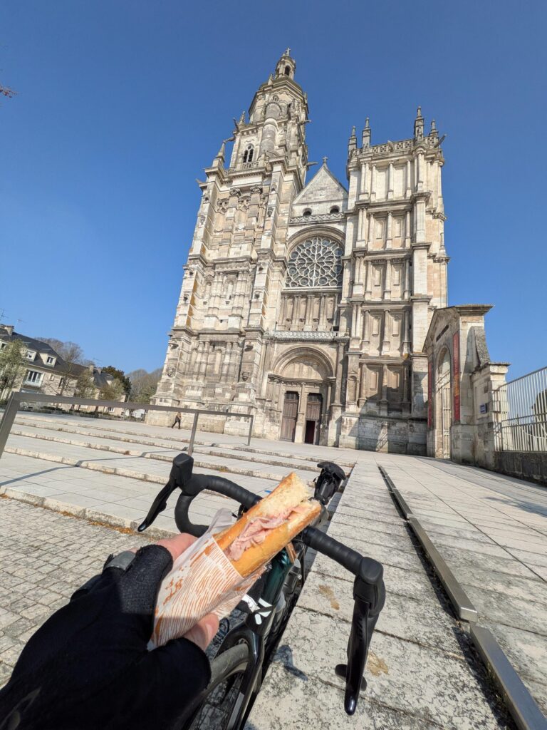 Vue subjective à la première personne d'un cycliste qui tient un sandwich à la main, devant son vélo. Au fond la cathédrale d'Évreux