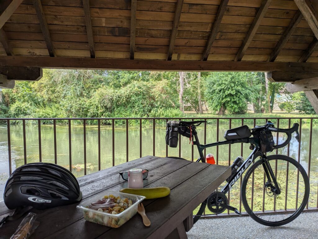 Sous un ancien lavoir, au premier plan une table de picnic en bois avec un yaourt une banane et une salle en plastique, derrière un vélo de course posé sur une barrière et derrière la rivière