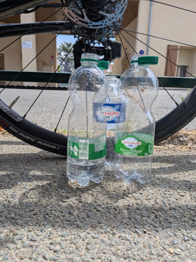 3 petites bouteilles d'eau vides en gros plan posées au sol devant une roue arrière de vélo