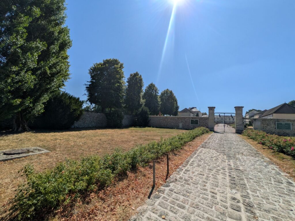 Une allée en pavés menant vers un château, à gauche une étendue d'herbe et un grand ciel bleu et soleil fort