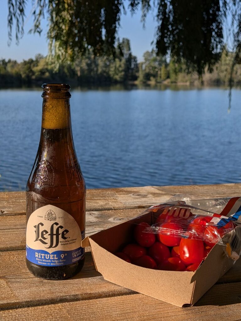 Une bouteille de bière et des tomates cerises posés sur une table en bois devant un lac au couché de soleil