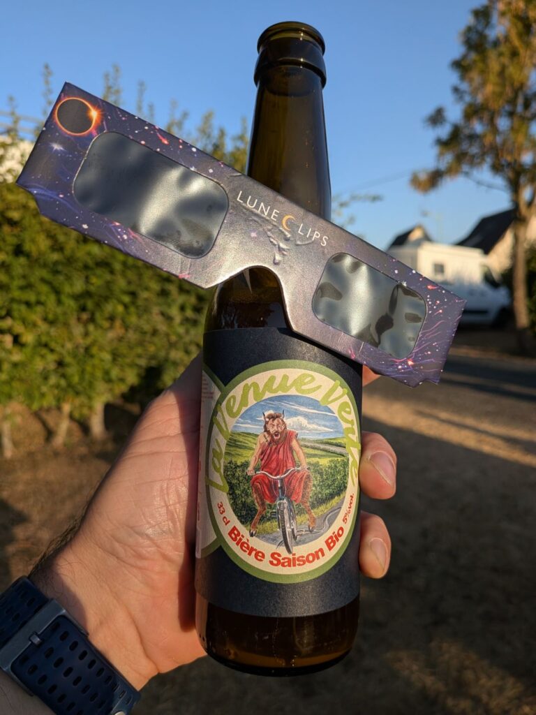 Une bouteille de bière "La Venue Verte" décorée d'un diable/mouton habillé de rouge faisant du vélo. Des lunettes d'observation du soleil pour éclipse sont posées sur la bouteille