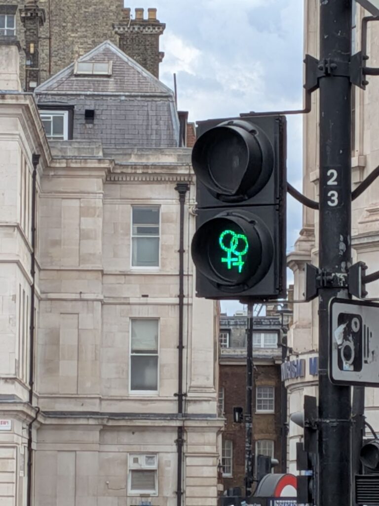 Un feu tricolore pour piéton dont le bonhomme vert a été remplacé par deux symboles féminins (cercle avec une croix en dessus) posées partiellement l'un sur l'autre