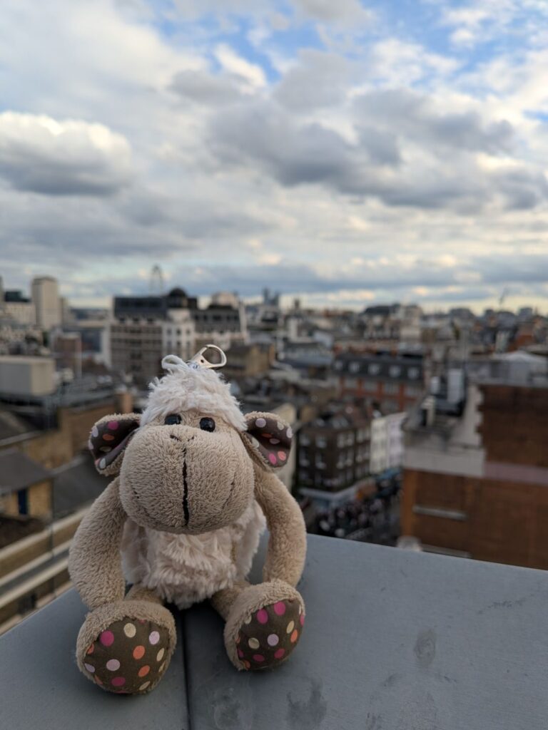 Posée sur un muret d'un toit une petit mouton en peluche, à l'arrière plan les toits de Londres