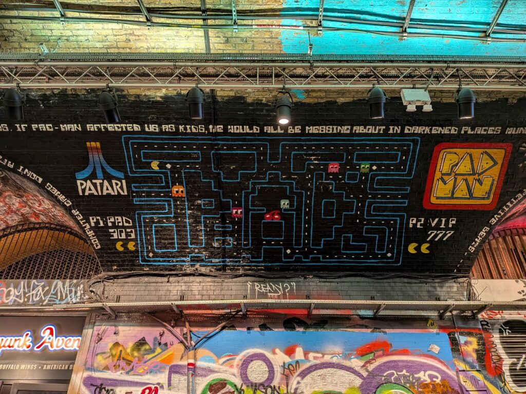 Un graph au plafond d'un tunnel représente une version du jeu PacMan mais renommé Padman de Patari. Texte "If PAC-MAN affected us as kids, the world will be messing about in darkened places running..." [after colored ghost]