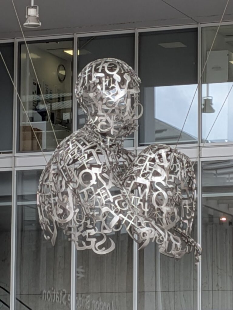 Sculpture suspendue d'un personnage vu de profil assis et tenant ses genoux dans les bras. Le bas du corps sous le torse n'est pas présent. La sculpture est en métal et constituée d'un ensemble de lettres formant le corps en 3 dimensions