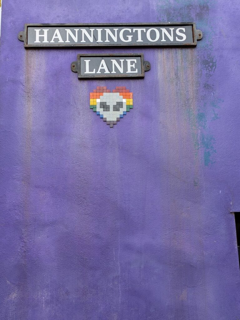 sur un mur violet, un panneau de rue (Hanningtons Lane) et dessous une mosaïque d'un alien sur un fond de cœur LGBT)