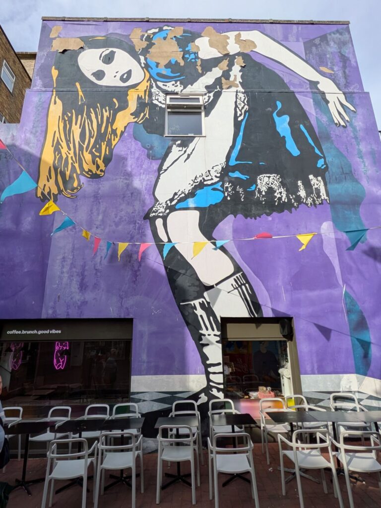 Un grand graph sur un mur peint en violet d'une personne pouvant être le personnage de Alice au pays des merveilles (blondes, robe courte bleue et tablier blanc) qui se tient penchée car elle est beaucoup plus grande que le mur