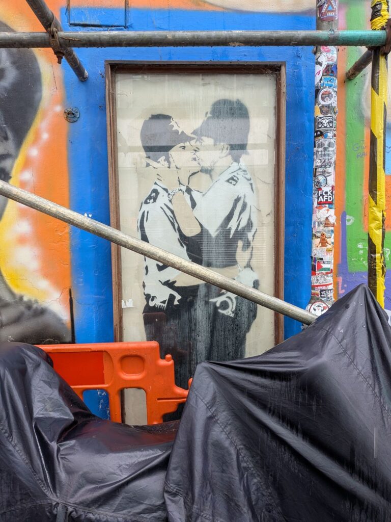 œuvre de street art de Banksy représentant deux policiers anglais qui s'enlacent et s'embrassent. L'œuvre est partiellement masquée par un échafaudage et un scooter sous une bâche