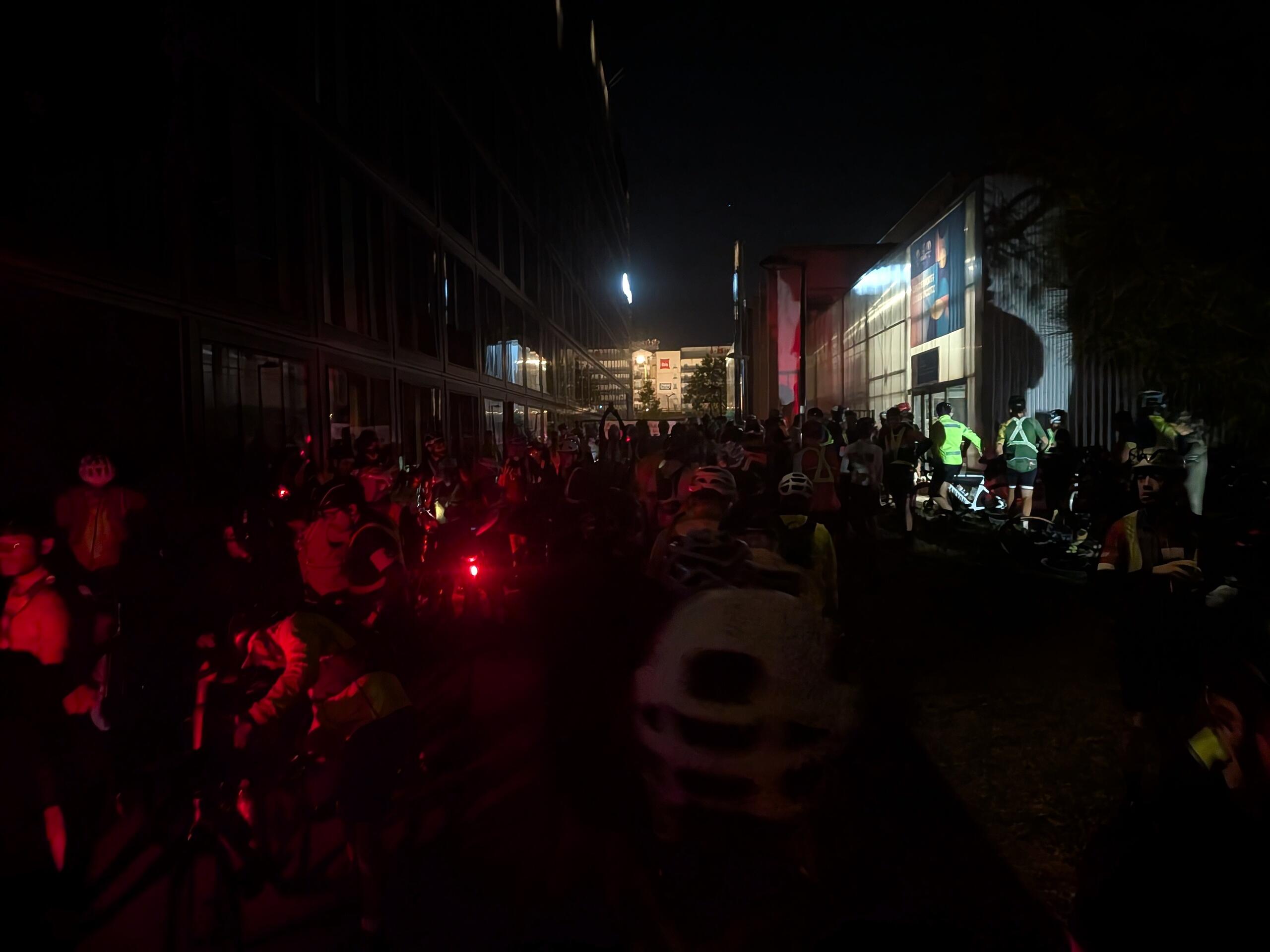 Plein de cyclistes de nuit très mal éclairé par des lumières rouges et blanches de vélo devant un gymnase