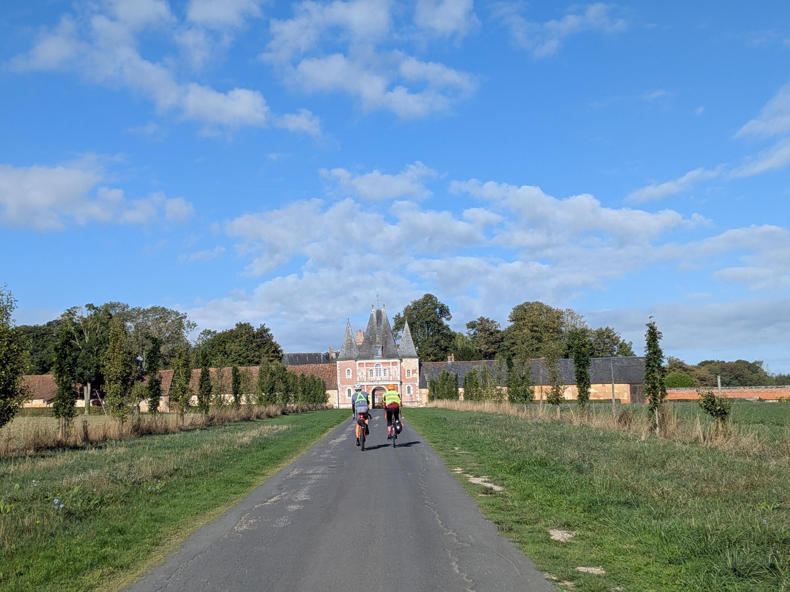 Deux cyclistes vus de dos sur une petite route en arrière plan l'entrée d'un château avec 3 tours en ardoise, mur roses