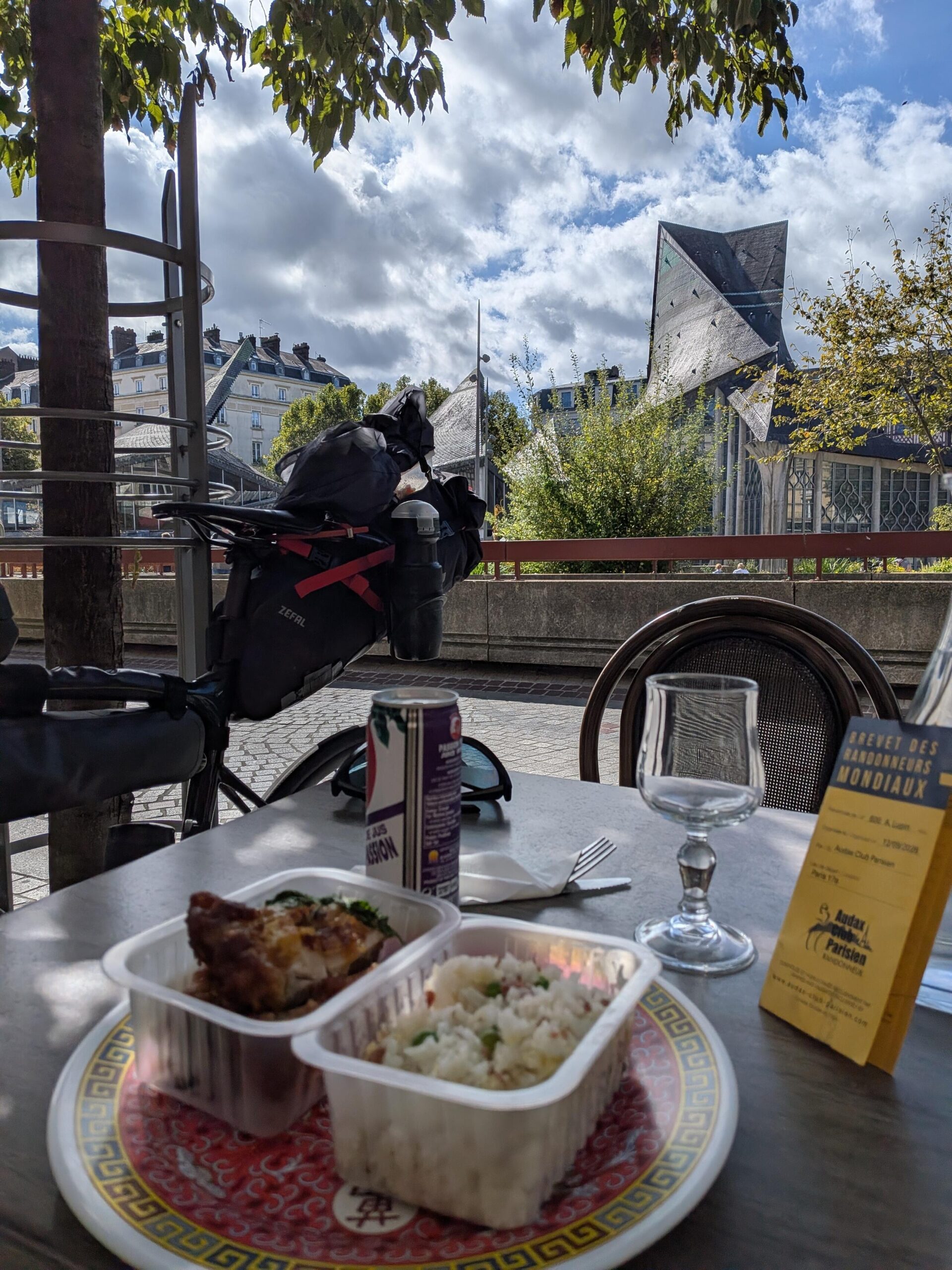 Un plat de nourriture asiatique sur une table en terrasse, un vélo en arrière plan et un petit papier jaune plié indiquant Brevet des randonneurs mondiaux