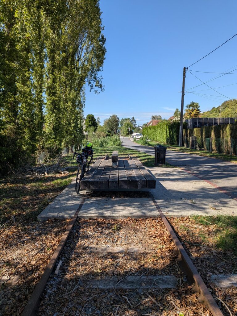 Sur une voie verte, le reste d'une voie de chemin de fer et un banc en bois large comme la voie sur lequel est posé un vélo