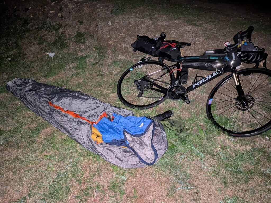 Depuis au flash, un vélo couché dans l'herbe et un campement de couchage simple avec bivy contenant matelas et duvet