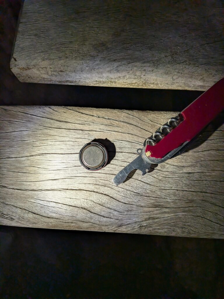 De nuit éclairé à la frontale sur un banc en bois une pile bouton et un couteau suisse ouvert