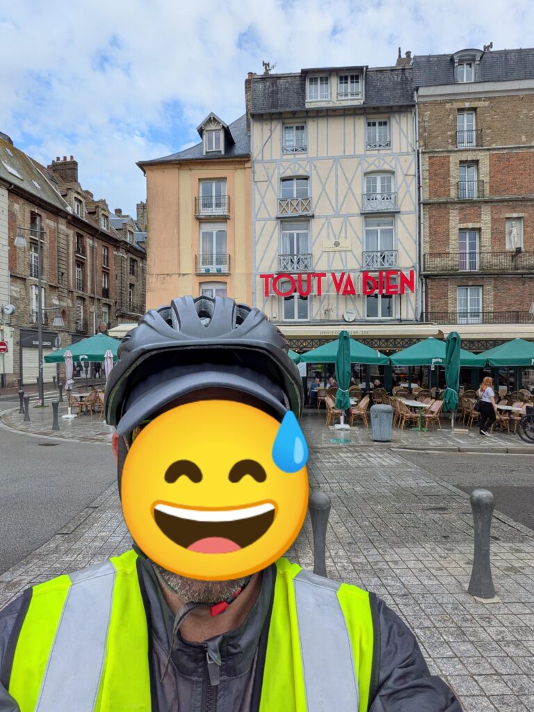 Un autoportrait d'une personne devant un café nommé "Tout va bien" (visage masqué par un smiley transpirant)