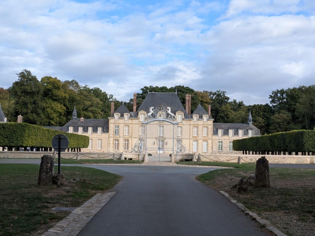 Vu d'un château tout blanc sur 2 étages plus combes, depuis une allée et devant grilles avec deux cheveaux